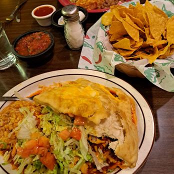 El Bruno's - Our lunch was delicious! - A plate of Mexican cuisine includes a burrito with a partially eaten tortilla, shredded lettuce, diced tomatoes, and beans. The meal is served on a ceramic plate with a side of rice and salad. In the background, there is a basket of tortilla chips, a small bowl of salsa, and condiments, along with additional dishes on the table. The setting appears to be a casual restaurant with a wooden table. - Mexican Near Me - Albuquerque, New Mexico
