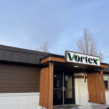 Vortex Eatery Plus Taphouse - Frontage - The image depicts the storefront of Vortex Eatery Plus Taphouse in Salem, Oregon. The building features a modern design with a combination of dark brown siding, white brick, and wooden accents around the entrance. The business sign displays the name 'Vortex' with a stylized logo. The entrance has glass doors, and the building's address, 1435, is visible on a vertical wooden panel. The sky above is clear with a few trees in the background. - Burgers Near Me - Salem, Oregon