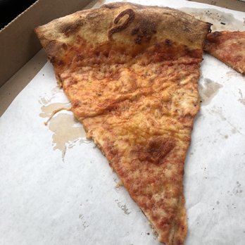 Nonna's Pizzeria - A large slice of cheese pizza with a thick, slightly browned crust and melted cheese is placed in a cardboard pizza box. The pizza has a golden-brown edge and a layer of melted cheese covering the surface. The box liner paper shows grease stains and some residual cheese. The image captures a typical takeout pizza serving. - Pizza Near Me - Patchogue, New York