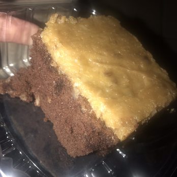Casa De Benavidez - A slice of cake with a yellow, possibly custard or fruit topping, is displayed inside a clear plastic container. The cake has a chocolate crust or base and appears moist. The image is taken at an angle, with a person's hand holding the container, and the background is dark and out of focus. - Mexican Near Me - Albuquerque, New Mexico