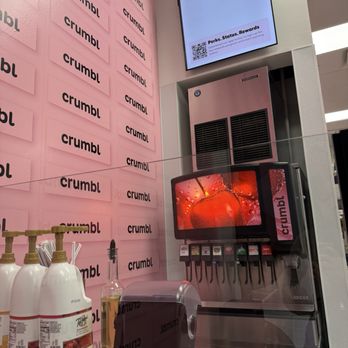 Crumbl - Orem - A drink station at Crumbl featuring a soda dispenser, bottles of syrup, and a pink wall with Crumbl logos, in a retail setting. - Bakeries Near Me - Orem, Utah