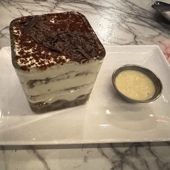 Piccolo Buco by Cooper's Hawk - A square tiramisu dessert featuring alternating layers of creamy mascarpone and coffee-soaked sponge cake, topped with cocoa powder and chocolate shavings. It is presented on a white rectangular plate alongside a small dish of custard or sauce, placed on a marble table surface. - Italian Near Me - Naperville, Illinois