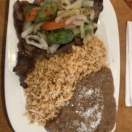 Bistec a La Mexicana - A rectangular white plate holds Mexican cuisine including rice, refried beans with cheese, grilled meat topped with sautéed vegetables, and a lemon wedge.