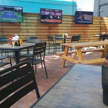 Laguna Burger - Nice outdoor patio - The image shows an outdoor dining area with multiple tables and chairs, including a picnic table, set on a brick-paved surface. A wooden privacy fence runs along the back, with three flat-screen TVs mounted on it, displaying tennis and basketball games. There are condiment bottles and napkin dispensers on some tables, and a person is seated on the right side. The setting appears to be a casual outdoor space for watching sports. - Burgers Near Me - Albuquerque, New Mexico