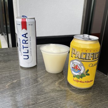  Mitchell's Mex - The image shows a wooden table with three items: a tall can of Michelob Ultra beer, a shorter can of Pacifico beer, and a clear plastic cup filled with a pale yellow beverage. The cans are positioned on either side of the cup, which appears to contain a frothy or cloudy drink. The background includes a white wall and part of a window or glass door, indicating an indoor setting. - Tex-Mex Near Me - Lafayette, Indiana