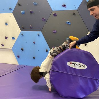 Over the Moon - Space space space - A child is lying on a purple padded mat, rolling over with their head down and legs up, while an adult holds a large purple crash pad labeled 'RESILITE' nearby. The setting is an indoor climbing gym with a wall covered in hexagonal panels painted in shades of blue, gray, black, and white, equipped with various climbing holds. The adult appears to be assisting or supervising the child during climbing activities. - Indoor Playcenter Near Me - Cary, North Carolina