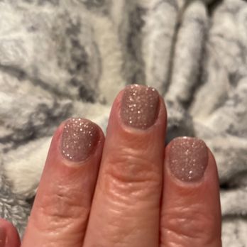 HERA Nails & Spa - The image features a close-up of three fingers with nails painted in a glittery, nude-colored polish. The nails are well-manicured, and the background appears to be a textured fabric with a gray and white pattern. The focus is on the nails, highlighting their shiny, sparkly finish. - Nail Salons Near Me - Warwick, Rhode Island