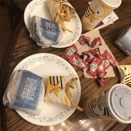 Double Cheeseburger Combo - The image shows two fast food meals on a wooden table, each with a burger wrapped in branded paper labeled 'Double Cheeseburger.' One burger has a side of French fries in a branded container, and there are multiple ketchup packets scattered on the table. Two large soft drinks in branded cups with straws are also present, along with a paper napkin. The setting appears to be a casual dining or takeout environment.
