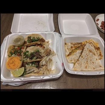 Mad Taco AZ - Tacos and quesadillas - Two foam takeout containers are placed on a wooden table, one containing tacos with shredded meat, onions, cilantro, and lime wedges, and the other holding folded quesadillas. Small containers of sauce and condiments are included. The image depicts a casual meal setting with disposable food containers and a partially visible plate of sliced meat in the background. - Food Trucks Near Me - Apache Junction, Arizona