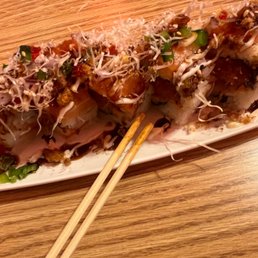 Red Dragon Rolls - A sushi roll is presented on a white oval plate placed on a wooden table. The roll is topped with sprouts, drizzled sauces, and chopped green onions. Chopsticks are positioned on the plate, indicating it is ready to be eaten. The setting suggests a Japanese or sushi restaurant environment.