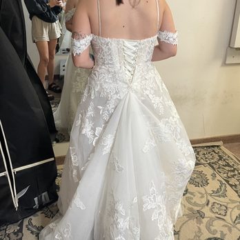 LD Alterations - My dress after (with bustle) - A woman is dressed in a white wedding gown with floral lace embroidery and off-shoulder lace sleeves. The dress features a lace-up back with satin ribbons, and the skirt has a layered, flowing design with embroidered floral patterns. The setting appears to be a fitting room or bridal shop, with other people visible in the background, some taking photos. - Sewing & Alterations Near Me - Indianapolis, Indiana