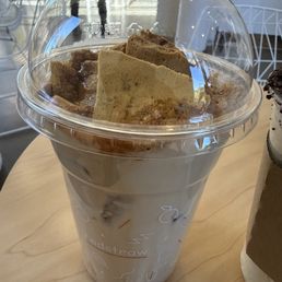 Dalgona cold brew coffee - A cold brew coffee beverage in a clear plastic cup with a domed lid, topped with foam and large biscuit pieces. The cup is on a light wooden table inside a cafe with modern decor, including white chairs and tables in the background.