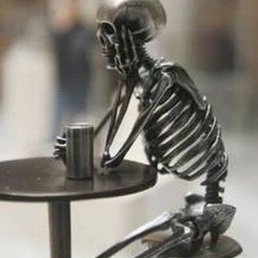 Me waiting for spicy mango wings... - A metallic sculpture of a skeleton is positioned at a small table, with one hand supporting its head and the other holding a can. The sculpture has a shiny, reflective surface and is seated on a stool, creating an artistic and contemplative scene.