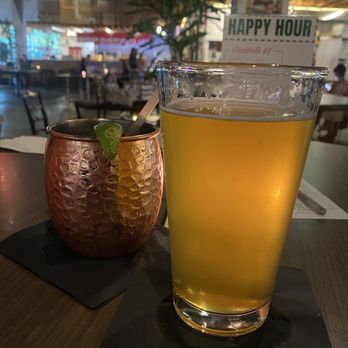 The Citadel - Mule & Beer from downstairs bar - A copper-colored cocktail with a lime wedge and a straw is positioned on a black napkin on a wooden table. Next to it is a tall glass of light-colored beer or cider. The background shows a busy indoor space with tables, chairs, and a sign that reads 'HAPPY HOUR.' The setting appears to be a food hall or bar area with ambient lighting and a casual atmosphere. - Food Court Near Me - Miami, Florida