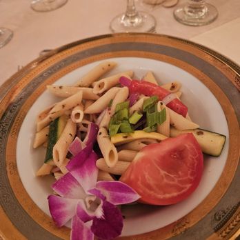 The Haley Mansion - A pasta salad featuring penne pasta mixed with chopped vegetables including tomato, cucumber, red onion, and green onion is presented on a decorative plate with a gold and black rim. An orchid flower is placed as a garnish on the side of the dish. The background shows a table setting with glasses and additional dishes, indicating a formal dining environment. - Party & Event Planning Near Me - Joliet, Illinois
