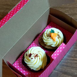 Red Velvet Cupcake - Two decorated cupcakes with frosting and toppings are inside a pink polka dot bakery box on a wooden surface.