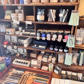 Boston General Store - A wooden shelving unit holds ink bottles, pens, notebooks, and stationery items, with a table of pens and writing instruments in front. - Accessories Near Me - Brookline, Massachusetts