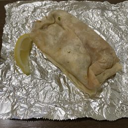 A burrito wrapped in a soft flour tortilla is resting on crinkled aluminum foil, with a lemon wedge placed nearby. The setting suggests it is from a Mexican restaurant or takeout service, with the foil used for serving or packaging. The background shows a dark surface, possibly a table or counter.