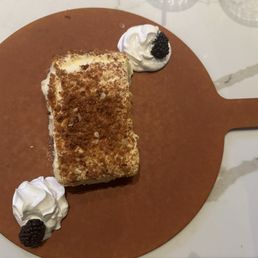 Almond Cake - A rectangular almond cake with a crumbly topping is placed on a round wooden serving board. The cake is accompanied by two dollops of whipped cream, each topped with a blackberry. The presentation is on a white marble surface, highlighting the dessert's textures and garnishes.