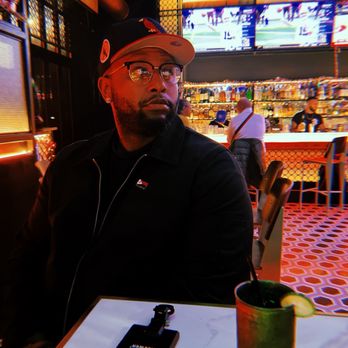 Dive Bar BK - A man with glasses and a cap sits at a bar, with a drink and a phone on the table, in a lively bar setting with multiple screens and bartenders. - Gastropubs Near Me - Brooklyn, New York