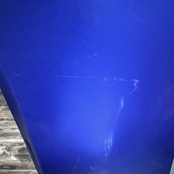 Cell ER - The image depicts a blue surface with multiple visible scratches and scuff marks. The surface appears to be made of plastic or painted material, with some areas showing more wear than others. The background includes a portion of a textured surface, possibly wood or laminate, and a small section of a greenish textured material at the bottom right corner. - Mobile Phone Repair Near Me - Spring, Texas