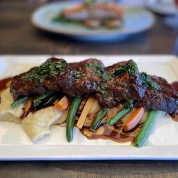 Short Ribs - A dish featuring short ribs with sauce, served over vegetables and mashed potatoes on a white rectangular plate.
