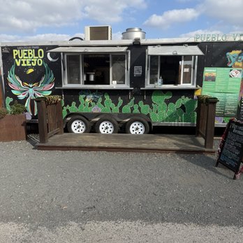 Meanwhile Brewing - A black food truck decorated with colorful cactus and butterfly artwork is parked outdoors under a partly cloudy sky. The truck has two service windows, one of which is open, with a QR code displayed nearby. There are three white wheels visible beneath the truck, and a small wooden platform in front of it. Menu boards are positioned on the right side, providing information about food options. - Breweries Near Me - Austin, Texas