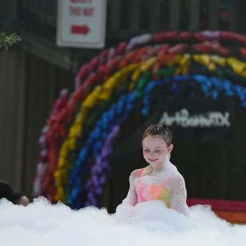 Art Barn ATX - A girl is enjoying herself in a foam or bubble-filled area outdoors, with a rainbow-colored arch and a sign in the background. - Art Classes Near Me - Leander, Texas