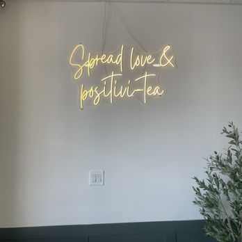 Tea Time - A neon sign with cursive writing displays the message 'Spread love & positivity-Tea' on a plain white wall, with a plant visible in the lower right corner. - Coffee & Tea Near Me - Albuquerque, New Mexico