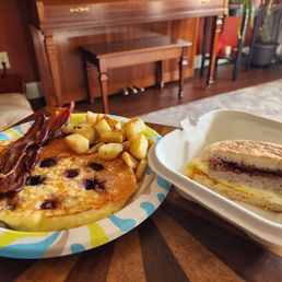 Blueberry pancake, bacon, home fries, and the Piermont sandwich. Chefs kiss! - A breakfast plate features a blueberry pancake, crispy bacon, home fries, and a sandwich in a small dish, set on a wooden table.