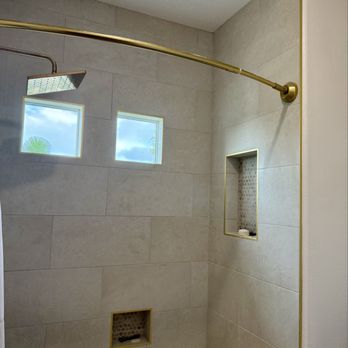 My Artistic Tile - Shower tile - A shower space with beige ceramic tiles on the walls, two small square windows providing natural light, and two recessed niches with patterned tile backgrounds for toiletries. A gold shower curtain rod extends across the space, and the showerhead is mounted on the wall. The design emphasizes clean lines and functional storage. - Tiling Near Me - Oxnard, California