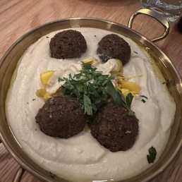 A metal bowl contains hummus topped with falafel balls, chopped herbs, and pickles, on a wooden table with a glass of water nearby.