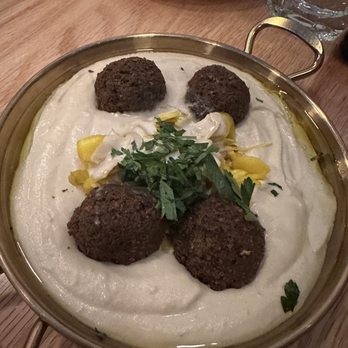 Levantine - A metal bowl contains hummus topped with falafel balls, chopped herbs, and pickles, on a wooden table with a glass of water nearby. - Mediterranean Near Me - Brooklyn, New York