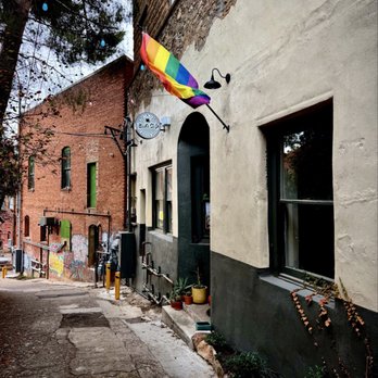 Earl's Cafe - The image shows a narrow alley with a building that has a beige and black exterior wall. A rainbow pride flag is mounted on the wall, fluttering in the breeze. The building has two windows and a door, with potted plants placed near the entrance. The alleyway has a rough, uneven pavement and is lined with other buildings, some with graffiti. Overhead, string lights are visible among trees, creating a colorful urban scene. - Coffee & Tea Near Me - Bisbee, Arizona