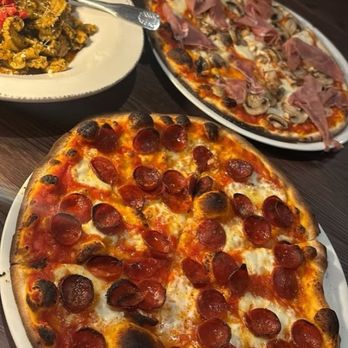 Piazza Caffe - Two pizzas, one topped with pepperoni and the other with ham and mushrooms, are on a dark wooden table. A pasta dish with red sauce and a spoon is also visible, along with wine glasses. - Tuscan Near Me - Santa Fe, New Mexico