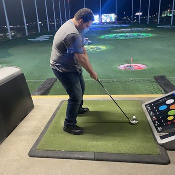 Topgolf - The view from the dining table on the 2nd floor tee box 1/3 - A man in a gray shirt and jeans, wearing a face mask, is standing on a green mat at an indoor mini golf course, holding a golf club. - Sports Bars Near Me - Glendale, Arizona