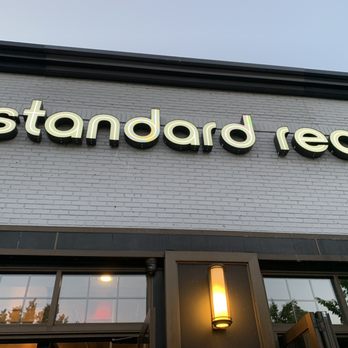 Standard Rec - The image depicts the exterior of Standard Rec, a nostalgic entertainment venue. The sign with the business name is mounted on a white brick wall, with illuminated lowercase letters spelling 'standard rec.' Below the sign, there is a window with reflections of trees and a warm exterior light fixture next to the entrance. The overall setting suggests a modern, inviting establishment. - Bars Near Me - Patchogue, New York