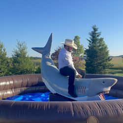Phantom Entertainment - A man in a white button down shirt and a beige cowboy hat, rides a large mechanical shark surrounded by a brown and blue inflatable ring.