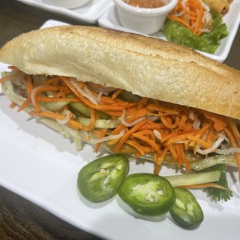 Pho Table - Bath Beach - Grilled Pork Banh Mi - A Vietnamese-style banh mi sandwich featuring grilled pork, shredded carrots, cucumbers, and pickled vegetables inside a baguette. The sandwich is presented on a white rectangular plate, garnished with sliced jalapeños and a sprig of cilantro. In the background, there are small bowls of dipping sauces and additional side salads, indicating a modern Vietnamese eatery setting. - Vietnamese Near Me - Brooklyn, New York
