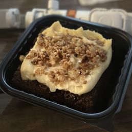 Chantilly Cake - A piece of chocolate cake topped with a layer of vanilla frosting and sprinkled with crushed nuts is presented in a black plastic container. The cake appears moist and rich, with the frosting and nuts adding texture. The background shows a wooden surface and electrical outlets, indicating an indoor setting, possibly a kitchen or dining area.