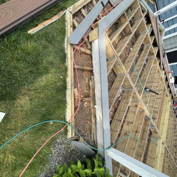 KnK Luxury Development - The image depicts a construction area where a wooden deck or platform is being built. The framing structure includes support beams, joists, and wooden boards, with some electrical wiring running across the site. Surrounding the construction are grass, plants, and building materials, indicating an outdoor setting. The construction appears to be in the early stages, with visible support structures and tools present. - Landscaping Near Me - Norwood, Massachusetts