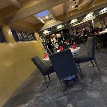 Amaya - The image shows an indoor restaurant space with a yellow painted wall and a ceiling with exposed wooden beams. The floor is made of large stone tiles. Several tables are set with red napkins, and black upholstered chairs surround them. In the background, patrons are seated, and staff members are attending to guests. The lighting is warm, and there is a decorative tapestry on the wall. The overall atmosphere appears to be casual and inviting. - New American Near Me - Santa Fe, New Mexico