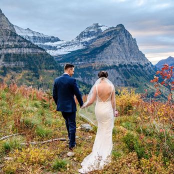 Bella Tailor - A bride and groom, dressed in wedding attire, walk hand-in-hand along a narrow mountain trail. The bride wears a white gown with an open back and a veil, while the groom is in a dark blue suit. The landscape features colorful autumn bushes and tall mountains with snow on their peaks under a partly cloudy sky. The scene captures a scenic outdoor wedding setting in a mountainous region. - Sewing & Alterations Near Me - Denver, Colorado