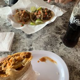 Chorizo Cheese Potatoes and Egg Burrito and Carnitas Taco - Three tacos with different toppings and lime wedges on a paper-lined tray, a burrito held in a hand, and a Coca-Cola bottle on a marble table.