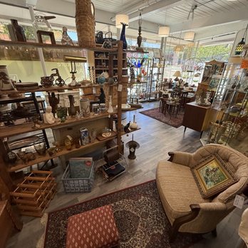 Vinteige - The image shows the interior of an eclectic vintage and antique store with wooden shelves and display units filled with decorative items, furniture, and lighting fixtures. A small upholstered armchair with a framed picture resting on it is positioned on a patterned rug in the foreground. The store has large windows at the back, allowing natural light to illuminate the space, and the ceiling features exposed pipes and hanging lamps. The overall setting is spacious and organized with a variety of vintage and antique pieces. - Furniture Stores Near Me - Walnut Creek, California