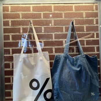 % Arabica - Two tote bags are displayed in a glass case mounted on a brick wall, one white with a black percentage symbol and one blue denim. - Coffee & Tea Near Me - Brooklyn, New York