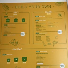 A yellow menu board offers customizable dairy-free acai bowls and smoothies, with options for bases, toppings, and add-ons.