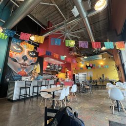 A Mexican restaurant interior featuring colorful papel picado banners, a large mural of a decorated sugar skull, and a casual dining setup with tables and chairs.