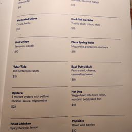 A menu page features a list of food and snack options including pretzel bites, marinated olives, oysters, fried chicken, and more. Each item has a brief description and price, with some items like oysters priced at $22 and others like marinated olives at $10. The menu is printed on a white background with blue and purple text, and the layout is organized into sections separated by lines.