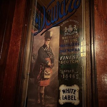 Ron's Place - Dewars in da house!! - A tall, rectangular mirror with a wooden frame displays an advertisement for Dewar's whisky, featuring a man dressed in traditional Scottish clothing, including a kilt and sporran. The mirror's surface includes text about Dewar's, its history, and branding, such as 'White Label' and 'Special Whiskies.' A paper towel roll is positioned at the bottom of the mirror, partially obscuring the lower part of the advertisement. The setting appears to be indoors with warm lighting and wooden wall paneling. - Pubs Near Me - Kenosha, Wisconsin